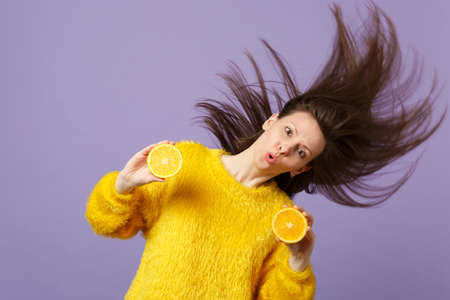 Amazed young woman in fur sweater with flowing hair holding halfs of fresh ripe orange fruit isolated on violet pastel background. People vivid lifestyle, relax vacation concept. Mock up copy spaceの写真素材