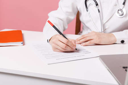 Cropped female doctor siting at desk working filling out medical documents in hospital isolated on pastel pink wall background indoors. Woman in medical gown stethoscope. Healthcare medicine conceptの写真素材