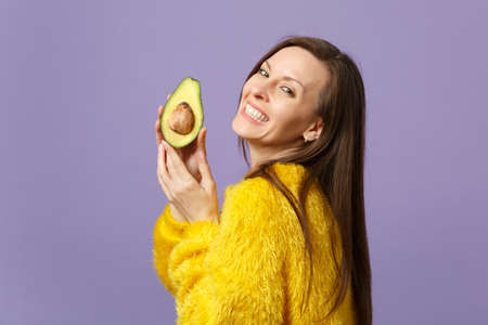 Smiling young woman in fur sweater holding in hand half of fresh ripe green avocado isolated on violet pastel background in studio. People vivid lifestyle, relax vacation concept. Mock up copy spaceの写真素材