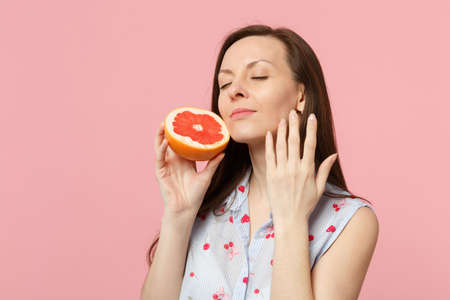 Beautiful young woman keeping eyes closed, put hand near face hold half of fresh ripe grapefruit isolated on pink pastel background. People vivid lifestyle relax vacation concept. Mock up copy spaceの写真素材