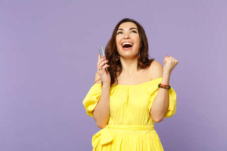 Happy young woman in summer dress using mobile phone, typing sms message, doing winner gesture, looking up isolated on violet background. People sincere emotions lifestyle concept. Mock up copy spaceの写真素材