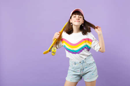Portrait of pretty teen girl in vivid clothes with yellow skateboard, holding hair isolated on violet pastel wall background in studio. People sincere emotions, lifestyle concept. Mock up copy spaceの写真素材