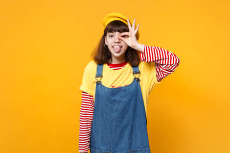 Funny girl teenager in french beret showing tongue holding hand near eye, imitating glasses or binoculars isolated on yellow background. People sincere emotions, lifestyle concept. Mock up copy spaceの写真素材