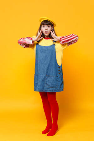 Full length portrait of shocked girl teenager in french beret and denim sundress keeping hands near...