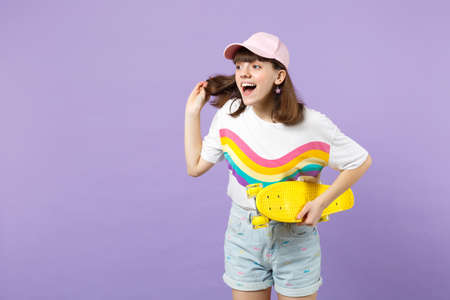 Excited teen girl in vivid clothes with skateboard holding hair, looking aside, keeping mouth open isolated on violet pastel background. People sincere emotions, lifestyle concept. Mock up copy spaceの写真素材