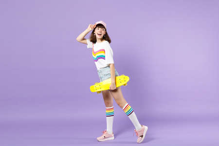 Side view of cheerful teen girl in vivid clothes holding yellow skateboard, looking back isolated on violet pastel background in studio. People sincere emotions, lifestyle concept. Mock up copy spaceの写真素材