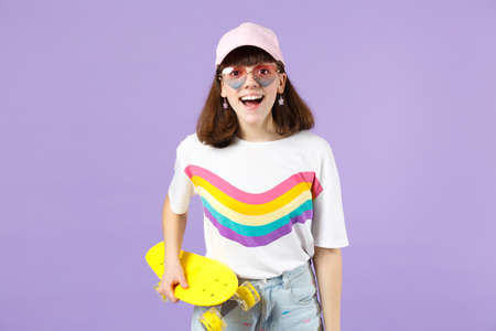 Excited teen girl in vivid clothes, heart eyeglasses holding yellow skateboard, keeping mouth open isolated on violet pastel background. People sincere emotions, lifestyle concept. Mock up copy spaceの写真素材