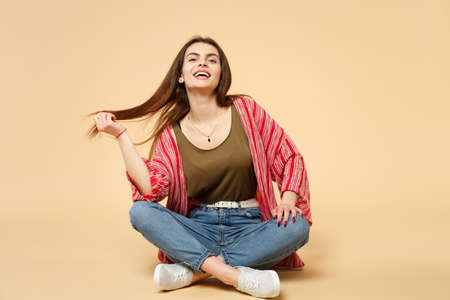 Portrait of laughing young woman in casual clothes sitting, looking camera, holding hair isolated on pastel beige background in studio. People sincere emotions, lifestyle concept. Mock up copy spaceの写真素材