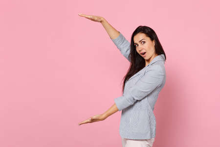 Side view of amazed young woman in striped jacket gesturing demonstrating size with vertical workspace isolated on pink pastel background. People sincere emotion lifestyle concept. Mock up copy spaceの写真素材