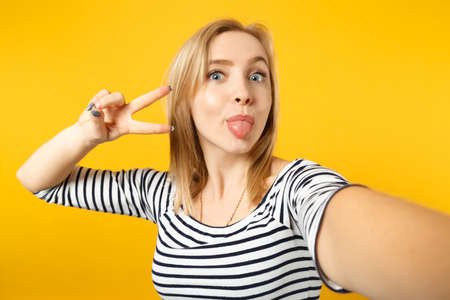 Close up selfie shot of funny young woman in striped clothes showing victory sign isolated on yellow orange wall background in studio. People sincere emotions, lifestyle concept. Mock up copy spaceの写真素材