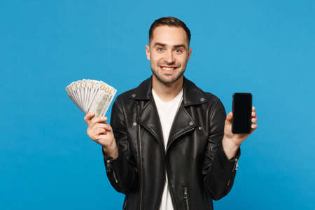 Young man in black jacket white t-shirt hold mobile phone empty screen for promotional content cash...