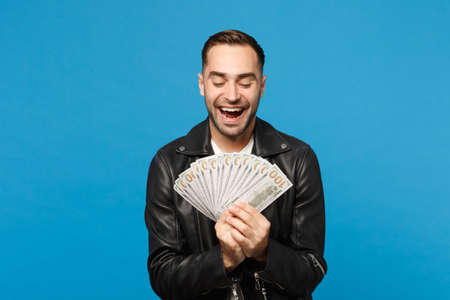 Young unshaven man in black leather jacket white t-shirt holding fan of cash money in dollar banknotes isolated on blue wall background studio portrait. People lifestyle concept. Mock up copy spaceの写真素材