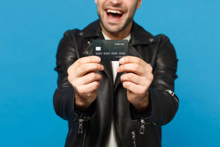 Close up stylish young unshaven man in black leather jacket white t-shirt hold in hand credit bank card isolated on blue wall background studio portrait. People lifestyle concept. Mock up copy spaceの写真素材