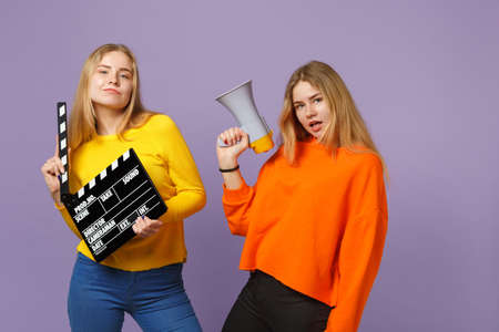 Two stunning young blonde twins sisters girls hold classic black film making clapperboard, scream on megaphone isolated on violet blue background. People family lifestyle concept. Mock up copy spaceの写真素材