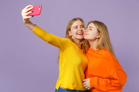 Two pretty young blonde twins sisters girls in colorful clothes doing selfie shot on mobile phone isolated on pastel violet blue wall background. People family lifestyle concept. Mock up copy spaceの写真素材