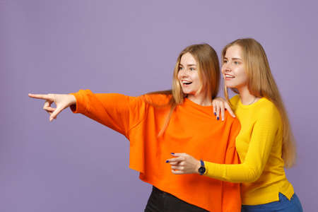 Two smiling young blonde twins sisters girls in vivid colorful clothes pointing index fingers aside isolated on pastel violet blue wall background. People family lifestyle concept. Mock up copy spaceの写真素材
