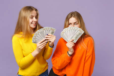 Two amazed blonde twins sisters girls in colorful clothes holding fan of money in dollar banknotes, cash money isolated on violet blue background. People family lifestyle concept. Mock up copy spaceの写真素材