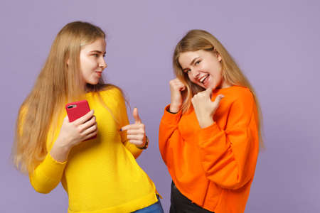 Two cheerful young blonde twins sisters girls in vivid clothes pointing thumb aside holding mobile phone isolated on pastel violet blue background. People family lifestyle concept. Mock up copy spaceの写真素材