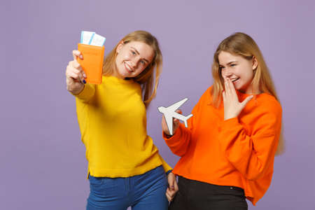 Two cheerful blonde twins sisters girls in colorful clothes hold passport, boarding pass ticket, paper airplane isolated on violet blue background. People family lifestyle concept. Mock up copy spaceの写真素材
