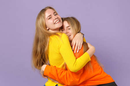 Two cheerful young blonde twins sisters girls in vivid colorful clothes hugging and looking camera isolated on pastel violet blue wall background. People family lifestyle concept. Mock up copy spaceの写真素材