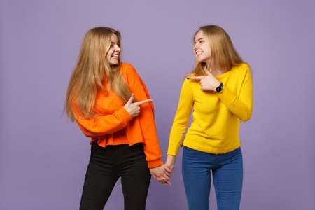 Two young blonde twins sisters girls in vivid clothes holding hands, pointing index finger at each other isolated on pastel violet blue background. People family lifestyle concept. Mock up copy spaceの写真素材