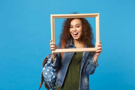 Young african american girl teen student in denim clothes, backpack hold frame isolated on blue background studio portrait. Education in high school university college concept. Mock up copy spaceの写真素材