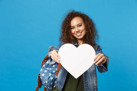 Young african american girl teen student in denim clothes, backpack hold heart isolated on blue...