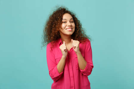 Portrait of smiling pretty african girl in pink casual clothes clenching fists isolated on blue turquoise wall background in studio. People sincere emotions, lifestyle concept. Mock up copy spaceの写真素材