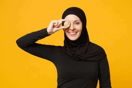 Young arabian muslim woman in hijab black clothes hold in hand holding bitcoin coin currency isolated on yellow background, studio portrait. People religious lifestyle concept. Mock up copy spaceの写真素材