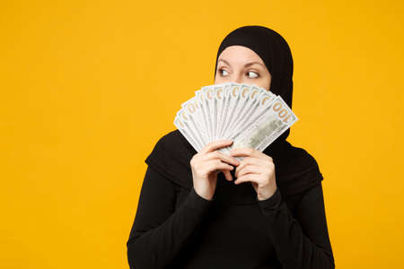 Young arabian muslim woman in hijab black clothes hold fan of cash money in dollar banknotes isolated on yellow wall background studio portrait. People religious lifestyle concept. Mock up copy spaceの写真素材