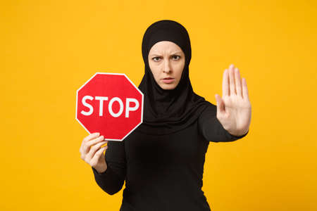 Young arabian muslim woman in hijab black clothes hold in hands red sign with Stop title isolated on yellow wall background, studio portrait. People religious lifestyle concept. Mock up copy spaceの写真素材