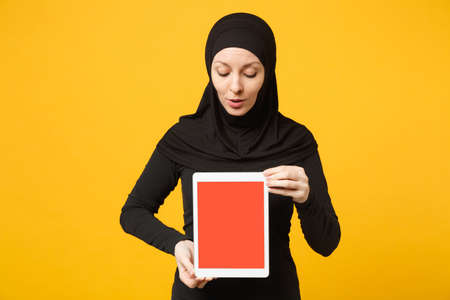 Young arabian muslim employee woman in hijab black clothes hold and work tablet pc computer isolated on yellow wall background, studio portrait. People religious lifestyle concept. Mock up copy spaceの写真素材