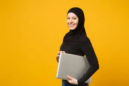 Young arabian muslim employee woman in hijab black clothes hold and work laptop pc computer isolated on yellow wall background, studio portrait. People religious lifestyle concept. Mock up copy spaceの写真素材