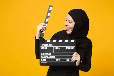Young arabian muslim woman in hijab black clothes hold classic black film making clapperboard isolated on yellow background studio portrait. People religious lifestyle concept. Mock up copy spaceの写真素材
