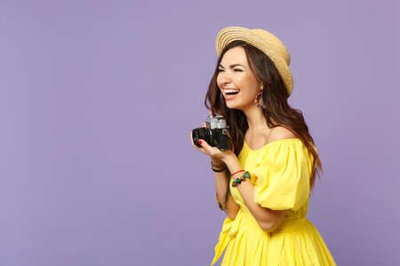 Laughing young woman in yellow dress hat taking picture on retro vintage photo camera looking aside...