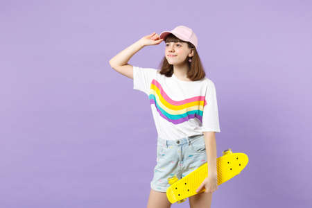 Portrait of charming teen girl in vivid clothes looking aside, holding yellow skateboard isolated on violet pastel background in studio. People sincere emotions, lifestyle concept. Mock up copy spaceの写真素材