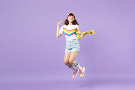 Pretty teen girl in vivid clothes holding yellow skateboard showing victory sign having fun jumping...