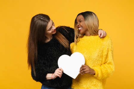 Two young women friends european and african american in black yellow clothes hold white heart isolated on bright orange wall background, studio portrait. People lifestyle concept. Mock up copy spaceの写真素材