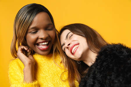 Two women friends european and african american in black yellow clothes doing selfie shot posing isolated on bright orange background studio portrait. People lifestyle concept. Mock up copy spaceの写真素材