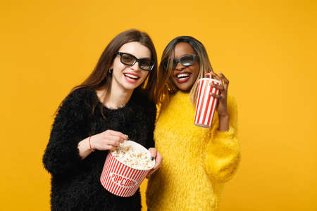Two women friends european and african american in black yellow clothes hold bucket of popcorn isolated on bright orange wall background, studio portrait. People lifestyle concept. Mock up copy spaceの写真素材
