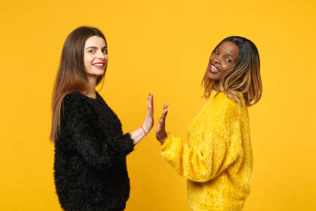 Two young women friends european and african american in black yellow clothes standing posing isolated on bright orange wall background, studio portrait. People lifestyle concept. Mock up copy spaceの写真素材
