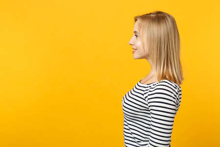 Side view of smiling attractive young woman in striped clothes stand, looking aside isolated on yellow orange wall background in studio. People sincere emotions, lifestyle concept. Mock up copy spaceの写真素材