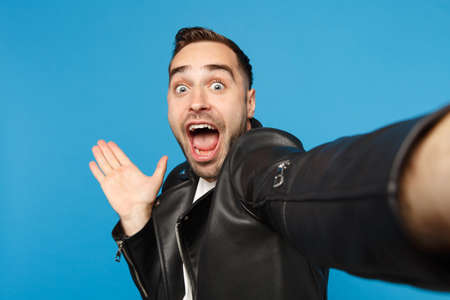 Close up selfie of stylish young unshaven man in black leather jacket white t-shirt looking camera isolated on blue background studio portrait. People sincere emotions concept. Mock up copy spaceの写真素材