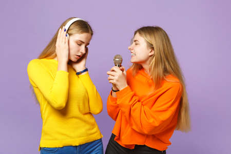 Two charming blonde twins sisters girls in vivid clothes listen music with headphones, sing song in microphone isolated on violet blue background. People family lifestyle concept. Mock up copy spaceの写真素材