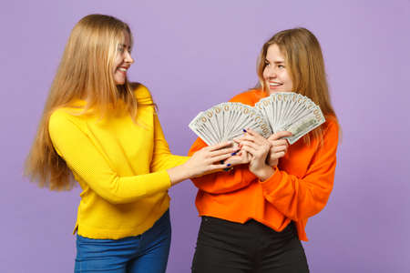 Two cheerful blonde twins sisters girls in colorful clothes holding fan of money in dollar banknotes cash money isolated on violet blue background. People family lifestyle concept. Mock up copy spaceの写真素材