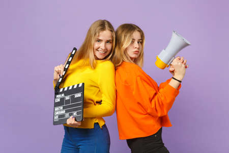Two smiling young blonde twins sisters girls holding classic black film making clapperboard scream on megaphone isolated on violet blue background. People family lifestyle concept. Mock up copy spaceの写真素材
