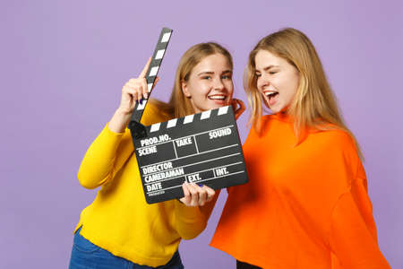 Two crazy young blonde twins sisters girls in colorful clothes holding classic black film making clapperboard isolated on violet blue background. People family lifestyle concept. Mock up copy spaceの写真素材