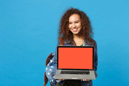 Young african american girl teen student in denim clothes, backpack work on pc isolated on blue background studio portrait. Education in high school university college concept. Mock up copy spaceの写真素材