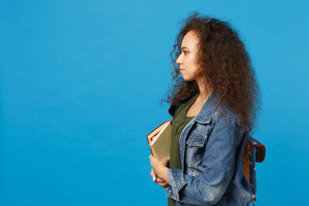 Young african american girl teen student in denim clothes, backpack hold books isolated on blue background studio portrait. Education in high school university college concept. Mock up copy spaceの写真素材