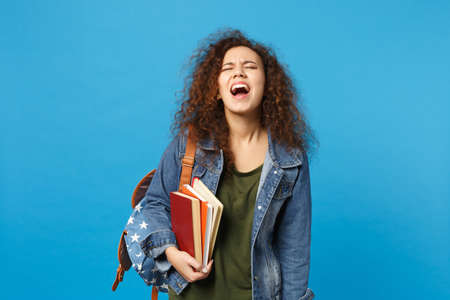Young sad african american girl teen student in denim clothes, backpack hold books isolated on blue background studio portrait. Education in high school university college concept. Mock up copy spaceの写真素材